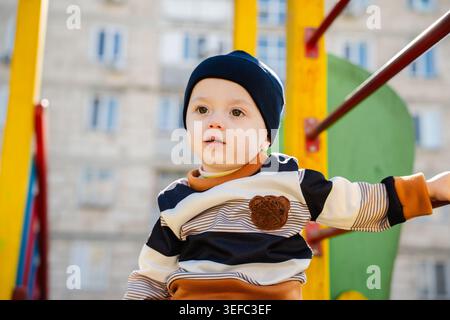 Bébé caucasien debout sur la structure de terrain de jeux dans la cour résidentielle ensoleillée. Enfance et mode de vie en plein air. Banque D'Images