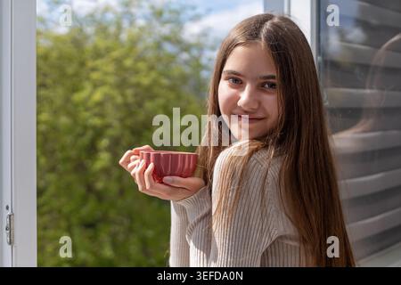 Sourire jeune femme buvant du thé par fenêtre. Jeune femme tenant une tasse de thé rouge par une fenêtre ouverte, souriant à la lumière naturelle avec des arbres verts à l'extérieur. Banque D'Images