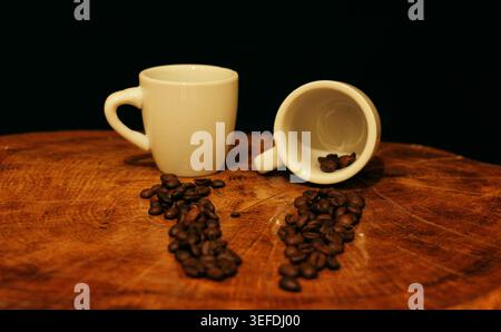 Grains de café torréfiés dispersés sur une surface en bois rustique à côté de deux tasses à expresso blanches, une verticale et une avec des grains renversés, backgroun foncé Banque D'Images