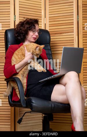 Femme dans un blazer rouge tient un chat au gingembre tout en utilisant un ordinateur portable dans un bureau à domicile, détendu et multitâche sur une chaise pivotante. Banque D'Images
