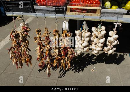 Suspendre des grappes de piments rouges séchés et de l'ail blanc à vendre à un étal de marché extérieur avec des tomates en arrière-plan Banque D'Images
