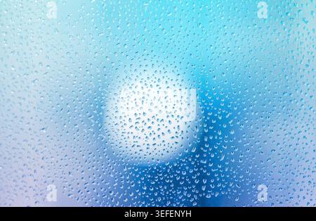 D'innombrables gouttelettes d'eau s'accrochent à une vitre, créant un motif texturé sur un fond bleu doux. Une source de lumière vive brille à travers le mouillé Banque D'Images