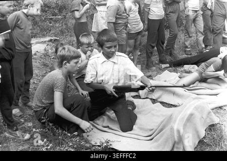 Sloviansk, 1983. Slavkurort, près du camp des pionniers de Vishnegorsk. Une image d'archives en noir et blanc en gros plan montrant deux garçons pendant une leçon de tir en plein air dans un ravin herbeux. Un garçon est agenouillé sur une couverture, tenant soigneusement et inspectant son fusil à air comprimé, tandis que son compagnon s'accroupit près de lui, regardant avec un intérêt intense. Cet instantané vintage détaillé souligne parfaitement la curiosité, la camaraderie et l'éducation sportive pratique des enfants soviétiques à l'époque historique d'un Donbass paisible. Banque D'Images
