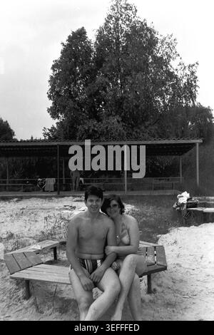 Sloviansk, 1983. Plage du camp pionnier de Vishnegorsk. Une image d’archives touchante en noir et blanc capturant Dmitry Mikhailovitch Kravelis et Inessa Mikhailovna Kravelis assis affectueusement ensemble sur un banc en bois. Ils sont vêtus de vêtements de plage, souriant joyeusement à la caméra avec un arbre feuillu massif et la structure d'ombre du camp en arrière-plan. Cet instantané franc du frère et de la sœur immortalise la joie, les liens familiaux et l'atmosphère relaxante des vacances d'été vécue à l'époque d'un Donbass paisible. Banque D'Images