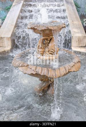 Fontaine en pierre détaillée avec deux niveaux avec de l'eau qui coule, entourée d'une piscine peu profonde, située dans un parc urbain avec une fresque colorée dans la BA Banque D'Images