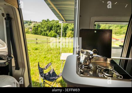 Vue intérieure d'une cuisine de camping-car moderne avec café infusé sur une cuisinière à gaz. La porte ouverte révèle une vue imprenable sur les collines verdoyantes Banque D'Images