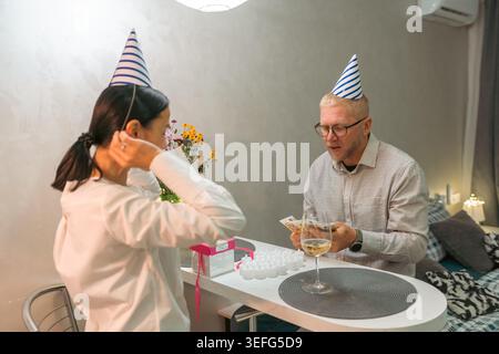 Chapeaux de fête couple célébrant l'anniversaire Appartement : célébration d'anniversaire intime ; un couple portant des chapeaux de fête profite d'un moment calme à la maison. Banque D'Images