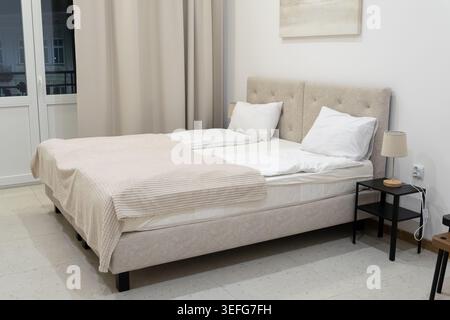 Une élégante chambre lumineuse arbore une tête de lit rembourrée avec des oreillers blancs et une couverture texturée, créant un sanctuaire paisible pour Mo Banque D'Images