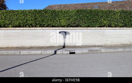 Clôture en mur de béton bordé par une rangée de briques et une haie sur le dessus. Longue ombre de lampadaire au milieu. Rooftop sur derrière, trottoir et rue Banque D'Images