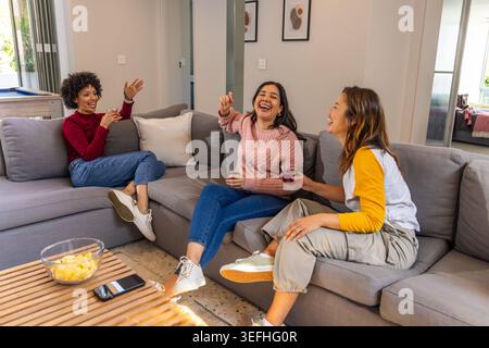 Diverses amies féminines riant et discutant sur le canapé sectionnel gris, tenant des boissons rouges Banque D'Images