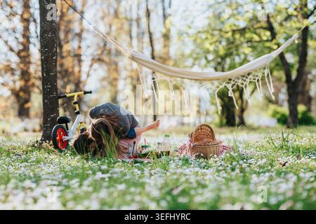 Mère avec des enfants jouant sur une couverture de pique-nique sous un hamac dans un parc ensoleillé Banque D'Images