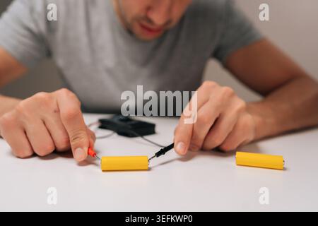 Homme effectuant un test de tension sur des batteries cylindriques jaunes avec un multimètre, connectant les sondes aux bornes de la table blanche, vérifiant la charge de la batterie Banque D'Images
