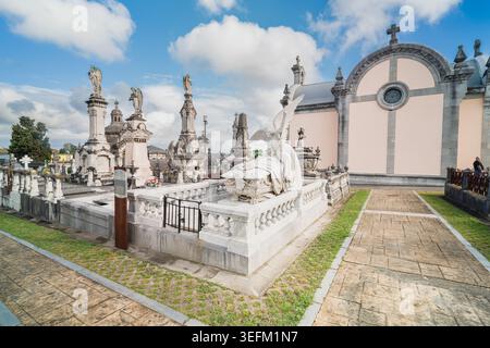 Vieux cimetière européen montrant de nombreux monuments funéraires ornés avec des statues d'ange, des pierres tombales et des croix, debout sous un ciel bleu à côté d'un grand c Banque D'Images