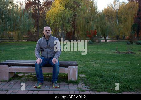 Homme d'âge moyen assis seul sur un banc de parc, portant une veste polaire grise et un Jean, regardant directement l'appareil photo avec un expert sérieux et pensif Banque D'Images