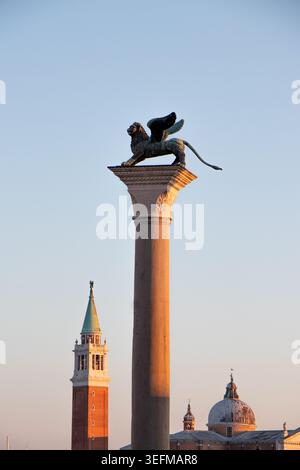 L'emblématique Lion ailé de Saint Marc se tenant au sommet d'une colonne historique au coucher du soleil à Venise, en Italie, avec le clocher et les dômes de San Giorgio Maggiore Banque D'Images
