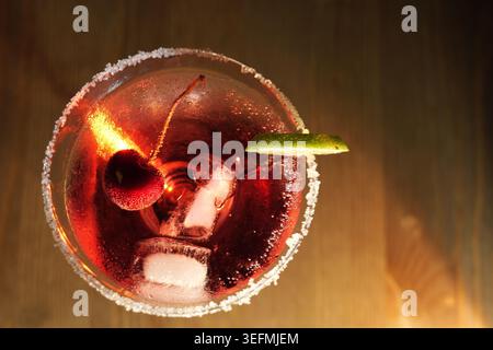 Cocktail rouge présentant des glaçons, une cerise et une tranche de citron vert, servi dans un verre à martini avec un bord de sel sur un fond rustique en bois, offrant un COO Banque D'Images