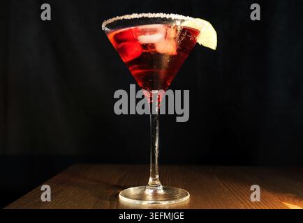 Cocktail de style martini rouge vif avec un bord de sel givré et un coin de citron vert, présenté sur un comptoir de bar en bois sur un fond sombre pour la vie nocturne A. Banque D'Images