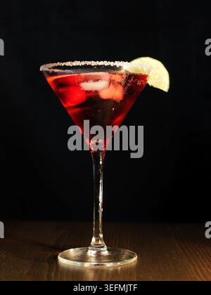 Cocktail rouge dans un verre à martini avec de la glace et un coin de citron vert, avec un bord sucré, reposant sur une surface en bois sombre avec un fond noir, capturin Banque D'Images