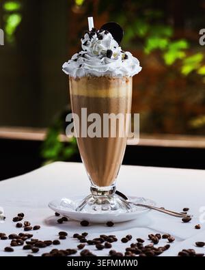 Milk-shake au café garni de crème fouettée et de copeaux de chocolat. Tourbillons de cacao dans un grand verre, servis sur soucoupe blanche. Grains de café dispersés autour de b Banque D'Images
