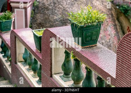 Des pots de fleurs en céramique émaillée verte se trouvent au sommet d'une rambarde en mosaïque rose au temple Kek Lok si à Penang, en Malaisie, avec d'énormes rochers de granit s'élevant derrière eux. Le contraste entre la céramique décorative et la pierre brute souligne le cadre naturel unique du temple. Banque D'Images