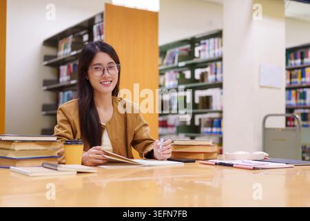 Jeune professionnelle asiatique étudiant avec des livres et des notes à la table de la bibliothèque souriante apprentissage des entreprises de recherche Banque D'Images