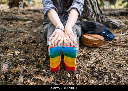 La personne est assise sur le sol de la forêt portant des chaussettes arc-en-ciel avec les pieds pointés vers la caméra. Représentation LGBTQ, vie queer authentique, personne non binaire Banque D'Images