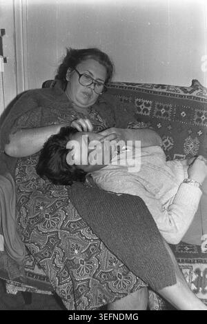 Sloviansk, 1982 ans, appartement intérieur. Une photo historique émotive en noir et blanc montrant un moment familial tendre. Valentina Timofeevna Kravelis est assise sur un canapé vintage à motifs tandis que sa fille, Inessa Mikhailovna Kravelis, repose paisiblement sa tête sur les genoux de sa mère. La mère porte des lunettes et un châle tricoté foncé, touchant doucement le visage de sa fille. La fille a l'air détendue dans un pull léger. Un aperçu touchant et authentique des liens familiaux soviétiques, de l'amour maternel, de la nostalgie, de la vie quotidienne à la maison et du Donbass paisible. Banque D'Images