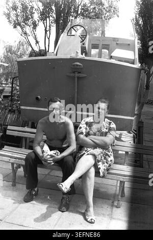 Sedove, 1982 ans, se reposant devant un bateau monté. Une photographie vintage en noir et blanc représentant un couple marié détendu en vacances. Kravelis Mikhail Ilyich et Kravelis Valentina Timofeevna sont assis sur un banc en bois, Valentina croisant les jambes et pliant confortablement les bras. Ils sont assis à l'ombre d'un grand bateau présenté comme une décoration de parc, profitant de l'ambiance calme de la station balnéaire. La photographie résume l'atmosphère d'une époque révolue et le souvenir de la vie avant la guerre. Banque D'Images