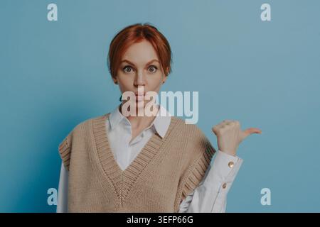 Femme de gingembre choquée dans des vêtements décontractés à la mode pointant avec le pouce à l'espace de copie, faisant l'expression du visage surpris, se sentant étonné par une bonne offre de vente ou une affaire, debout isolé sur fond bleu Banque D'Images