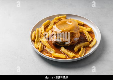 Gros plan d'un sandwich Francesinha portugais traditionnel recouvert de fromage fondu et d'une riche sauce tomate, entouré de frites dorées sur un whi Banque D'Images