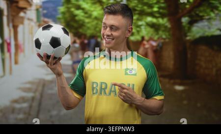 Man in brasil maillot tient le ballon de football avec la main droite et pointe le doigt tout en souriant dans la rue ; fierté joyeuse. Banque D'Images