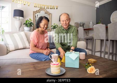 Femme asiatique senior et homme asiatique mature assis sur le canapé ouvrant la boîte cadeau sarcelle près du gâteau Banque D'Images