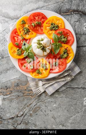 fromage burrata servi avec tranches de tomates rouges et jaunes, feuilles de basilic frais et sauce pesto vert sur une assiette blanche avec deux fourchettes sur un dos de pierre Banque D'Images