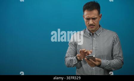 Homme tapant le smartphone avec l'index tout en tenant le téléphone dans les mains sur fond bleu dans le studio ; concentration de frustration. Banque D'Images