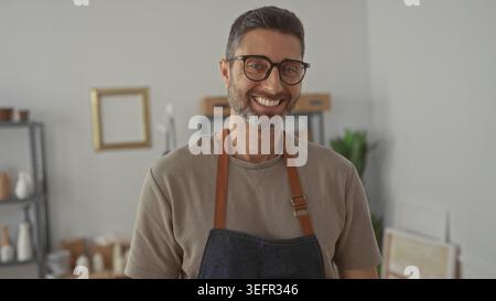 Homme souriant dans un tablier en denim parmi des pots en céramique sur des étagères en studio portant des lunettes et de la barbe, vue de face ; fierté créative. Banque D'Images