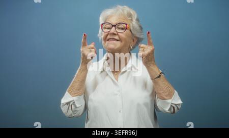 La femme senior fait des gestes avec les index sur ses joues puis pointe les deux index vers le haut, portant des lunettes colorées et un chemisier blanc en studio ; pla Banque D'Images