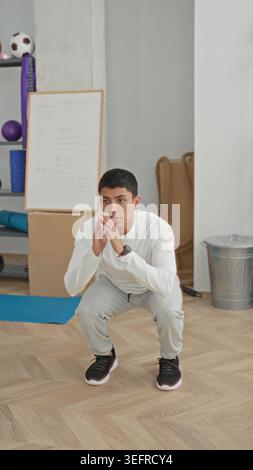 Homme effectuant un squat avec les mains attachées, portant des baskets noires et des pantalons de survêtement gris dans un studio d'entraînement avec tapis de yoga et tableau blanc ; concentration Banque D'Images