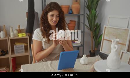 Femme portant tablier tient petit vase en céramique avec les mains et les gestes vers la tablette sur la table de travail en studio ; concentration créative. Banque D'Images