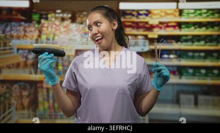 Femme dentiste tient guide de teinte de dent et sondes dentaires, souriant avec les mains gantées dans la construction de supermarché ; confiance ludique. Banque D'Images