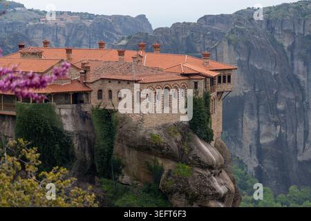 Vue détaillée des arches en pierre et des toits en terre cuite du monastère de Varlaam, avec des fleurs roses et le Grand Météoron visible au loin. Banque D'Images