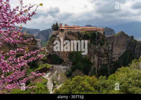 Une vue spectaculaire sur le monastère Saint de Varlaam encadré par un arbre Judas en fleurs, sur fond d'anciens piliers rocheux et d'un ciel nuageux. Banque D'Images