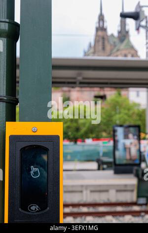 Un validateur de billet jaune vif monté sur un poteau à un arrêt de tramway urbain, avec des flèches floues de l'église, un abri de plate-forme et des pistes en arrière-plan co Banque D'Images