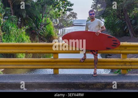 Homme athlétique en t-shirt et boardshort debout sur un pont routier jaune tenant une planche de surf rouge, végétation tropicale et rivière derrière lui, Ocean visi Banque D'Images