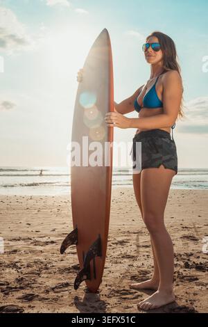 Jeune femme en bikini et short tenant une planche de surf tout en regardant vers l'horizon sur une plage ensoleillée. Scène rétro-éclairée avec reflets d'objectif et vagues océaniques Banque D'Images