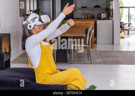 Dame senior dans la tenue jaune à l'aide d'un casque VR assis sur le siège à la maison à l'aide de smartwatch Banque D'Images