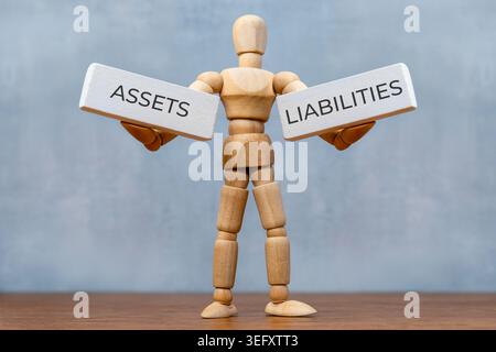 Un mannequin en bois tenant deux blocs blancs étiquetés ACTIFS et PASSIFS sur fond gris. Équilibre financier, comptabilité personnelle Banque D'Images