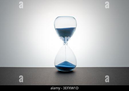 Un sablier en verre avec du sable bleu coulant repose sur une surface sombre sur un fond dégradé lumineux et rétroéclairé. Banque D'Images