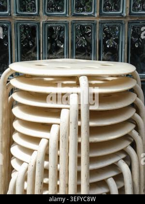 Gros plan de tabourets empilés en bois rond avec pieds courbés et grain de bois moucheté, disposés devant un mur de blocs de verre ensoleillé. Composition minimaliste avec R Banque D'Images