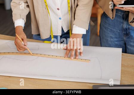 Deux créateurs de mode utilisent une règle incurvée et un crayon pour dessiner des patrons de vêtements sur un grand papier illustrant la conception collaborative et les compétences de couture. Banque D'Images