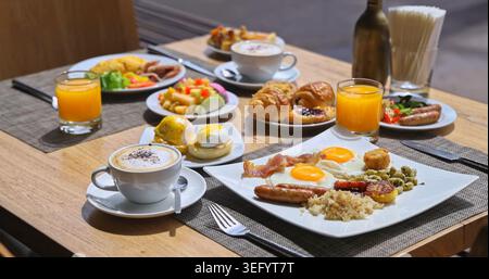 Plats de petit déjeuner assortis assis sur une table en bois dans un café ensoleillé, œufs frits, bacon, pâtisseries, fruits frais et café, lumière vive du matin, luxe chaud Banque D'Images
